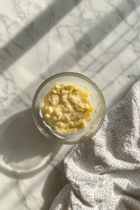 This image shows a small clear glass bowl on a white marble countertop containing Greek yogurt, mayonnaise, and lemon juice being combined into a creamy sauce.