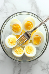 This image shows hard-boiled eggs being lightly mashed in a bowl using a fork, forming the base of a creamy egg salad.