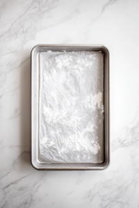 This image shows a silver jelly roll pan lined with parchment paper and lightly greased and floured on a white marble countertop.