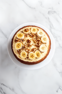This image shows the banana walnut cake being carefully assembled, with whipped cream between layers and a final topping of banana slices and chopped walnuts.