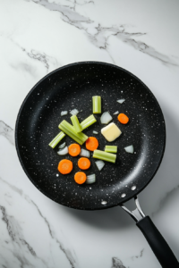 This image shows a black non-stick skillet on a white marble cooktop, melting butter and sautéing chopped onions, celery, carrots, and minced garlic over medium-high heat.