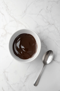 This image shows semi-melted chocolate in a white microwave-safe bowl placed on a spotless white marble cooktop. A silver spoon rests beside the bowl, and the glossy surface of the chocolate reflects natural lighting, creating a realistic and mouth-watering texture.