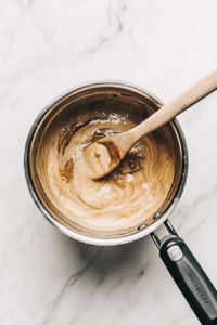 This image shows a shiny stainless steel saucepan placed on a white marble cooktop, with sugar, unsalted butter, and ¼ cup water beginning to melt together, and a wooden spoon resting nearby.