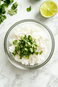 This image shows a clear glass bowl on a white marble surface with a well-mixed rice, cilantro, and lime juice mixture. The green herbs are evenly distributed throughout the white rice.
