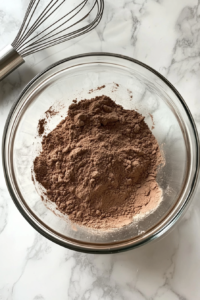 This image shows a top-down view of dry ingredients including sugar, flour, cocoa powder, baking powder, baking soda, and salt stirred together in a clear glass mixing bowl on a white marble countertop.