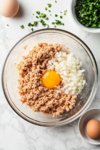 This image shows a glass bowl on a white marble surface containing ground chicken, breadcrumbs, chopped onion, egg, and minced garlic ready to be mixed.