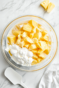 This image shows a large mixing bowl filled with pineapple chunks, sugar, cornstarch, water, eggs, and vanilla being whisked together into a smooth, golden mixture.