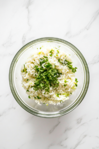 This image shows a clear glass bowl filled with cooked white rice, freshly chopped green cilantro, and lime juice, placed on a white marble countertop.