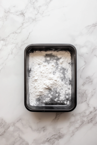 This image shows a top-down view of a black 9×13-inch nonstick baking dish sprayed with flour-based nonstick spray over a white marble surface.