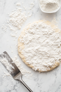 This image shows the risen dough patted into a 20 cm disc on a lightly floured white marble surface, with a metal dough scraper resting nearby.