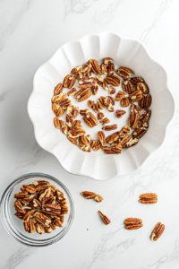 This image shows a top-down view of chopped pecans evenly spread inside a clean white 10-inch Bundt pan on a white marble cooktop, with a small glass bowl of extra pecans beside it.
