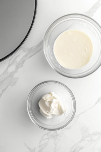 This image shows 4 cups of buttercream set aside in a small clear glass bowl next to the larger bowl, all placed over a white marble surface.