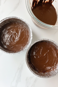 This image shows a top-down view of thin chocolate cake batter being poured evenly into two greased and floured baking pans placed on a white marble countertop.