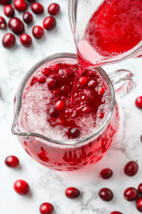 This image shows cranberry juice, pineapple juice, and fizzy ginger ale being poured into a tall pitcher, blending together to create the perfect fruity base for a party punch.