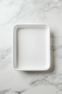 This image shows a preheated oven at 350°F with a white ceramic 9x13-inch baking dish lightly greased with butter, resting on a white marble cooktop.