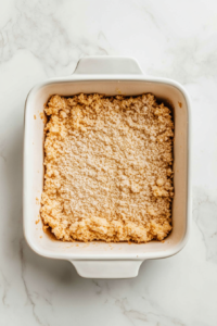 This image shows the cake mix dough being pressed evenly into the bottom of a greased ceramic baking dish using clean hands. The white marble countertop is visible beneath, with a clean background.