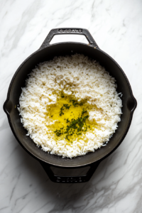 This image shows uncooked white rice, chicken broth, and olive oil added into a black skillet on a white marble cooktop.