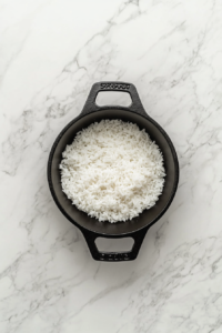 This image shows rice simmering gently in chicken broth and olive oil inside a black skillet placed on a white marble countertop.