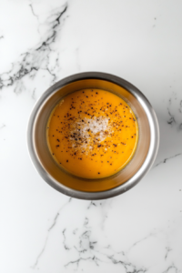 This image shows creamy butternut squash soup inside a stainless steel pot with freshly added salt and black pepper on top, placed on a white marble cooktop.