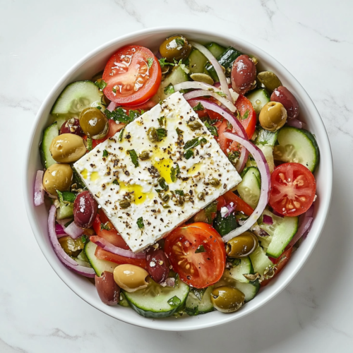 This image shows a vibrant Athens Salad served in a round white bowl, filled with chopped tomatoes, crisp cucumbers, and red onions, all topped generously with freshly ground black pepper.