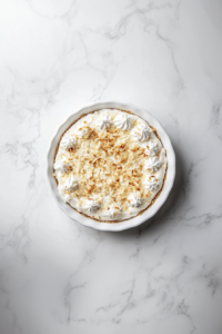 This image shows the chilled pina colada pie being topped with a cloud of whipped cream and a final scattering of coconut flakes for a dreamy tropical finish.