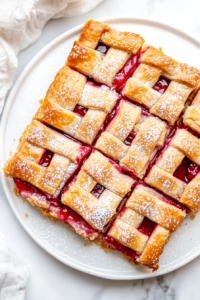 This image shows sliced cherry pie bars arranged on a white ceramic plate, with a sugar glaze drizzle and powdered sugar topping over golden criss-cross crusts, set on a clean white marble surface.