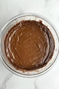 This image shows a top-down view of smooth and glossy chocolate cake batter in a clear glass mixing bowl after being beaten with an electric mixer, placed on a white marble countertop.