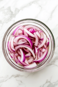 This image shows thinly sliced red onions soaking in a clear glass bowl filled with ice water and a splash of red wine vinegar. The bowl sits directly on a clean white marble countertop, with no background clutter.