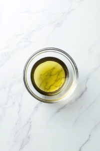 This image shows a clear glass mixing bowl placed on a white marble countertop containing a freshly whisked dressing made from garlic chili sauce, lime juice, brown sugar, and water.
