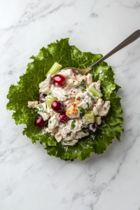 This image shows a spoonful of chicken salad mixture being placed in the center of a crisp lettuce leaf on a white marble countertop.