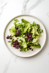 This image shows a simple white ceramic plate over a white marble countertop topped with a fresh layer of spring salad mix as a base for the chicken and mango topping.