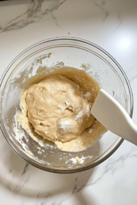 This image shows the sticky but structured dough resting inside a clear glass bowl, with a white rubber spatula beside it on a clean white marble cooktop.
