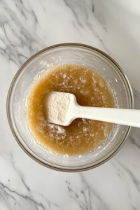 This image shows very warm water being stirred into the flour mixture using the handle of a white rubber spatula inside a clear glass bowl, all placed neatly on a white marble countertop.