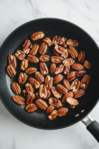This image shows pecan halves toasting in a large black nonstick skillet placed on a clean white marble cooktop.