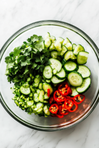 This image shows a colorful mix of sliced cucumber, creamy avocado, chopped scallions, chili pepper, and fresh cilantro in a medium bowl.