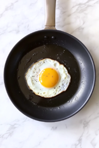 This image shows eggs cooking in a skillet of hot olive oil, seasoned with a sprinkle of salt and pepper for a savory morning touch.