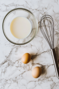This image shows a medium clear glass bowl with buttermilk, oil, eggs, and vanilla extract whisked together on a clean white marble countertop.