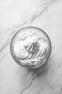 This image shows whipped cream beaten with sugar and vanilla in a clear glass bowl until stiff peaks form, resting on a white marble countertop.