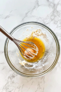 This image shows a large clear glass bowl on a white marble cooktop containing Greek yogurt, granulated sugar, eggs, olive oil, and vanilla extract, being whisked together until smooth.