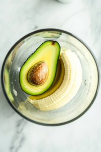 This image shows a clear glass blender jar sitting on a white marble countertop, containing one peeled avocado and half a peeled banana inside, ready to blend.