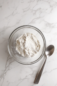 This image shows one cup of flour being mixed into the egg, milk, and salt mixture in a glass bowl over a white marble countertop.