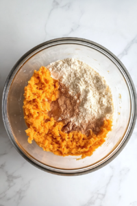 This image shows mashed sweet potato added to a glass mixing bowl filled with flour, cornmeal, sugar, baking powder, baking soda, salt, and cinnamon, all placed on a white marble countertop.