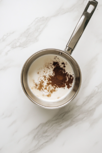 This image shows a shiny stainless steel saucepan on a white marble cooktop filled with whole milk, heavy cream, powdered sugar, and espresso powder, ready to heat.