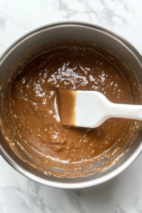 This image shows milk being added into a roux inside a stainless steel pot on a clean white marble cooktop, stirred with a white silicone spatula.