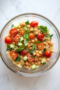This image shows a clear glass mixing bowl on a white marble countertop, filled with couscous, halved fresh cherry tomatoes, roasted cherry tomatoes, chickpeas, cucumber, feta, and basil before mixing.