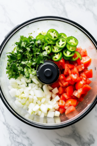 This image shows a clear food processor bowl filled with chopped onion, garlic, diced tomatoes, sliced jalapeños, fresh cilantro leaves, lime juice, lime zest, ground cumin, salt, and sugar. The equipment is on a white marble countertop.