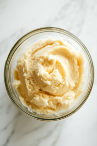 This image shows a top-down view of a glass bowl on a white marble countertop, with vanilla extract and flour added on top of a creamed butter and sugar mixture, ready to be mixed.