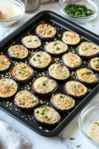 This image shows a black baking sheet with eggplant slices coated in breadcrumbs, arranged in a single layer on a white marble countertop.