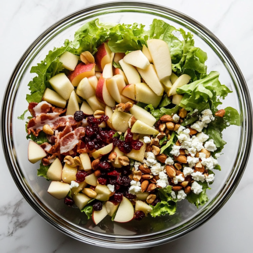 This image shows a top-down view of Autumn Chopped Salad in a large white ceramic bowl on a white marble countertop, featuring a colorful mix of fresh fruits, vegetables, bacon, and cheese.