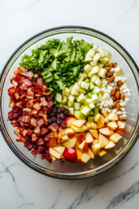 This image shows a large glass bowl on a white marble cooktop with chopped romaine lettuce, bacon, apples, pear, feta, peanuts, and cranberries layered inside.